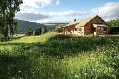 A wooden house in a lush green meadow with hills and trees under a partly cloudy sky.