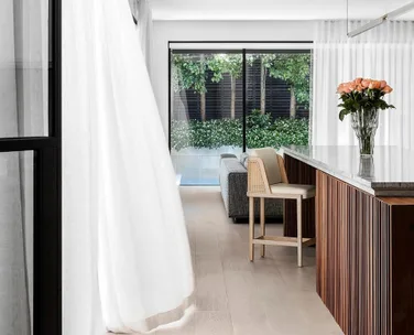 Modern kitchen with marble countertop, flowers in a vase, and large windows with sheer curtains opening to a garden view.