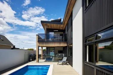 Modern house exterior with a pool, glass balcony, and lounge chair, set against a bright blue sky.