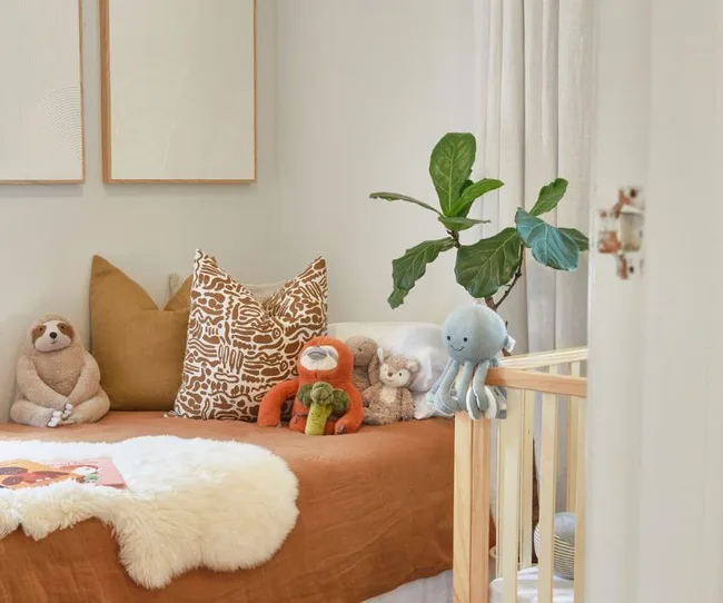 Cozy child&rsquo;s room with plush toys and pillows on a bed, soft colors, and a green plant next to a crib.
