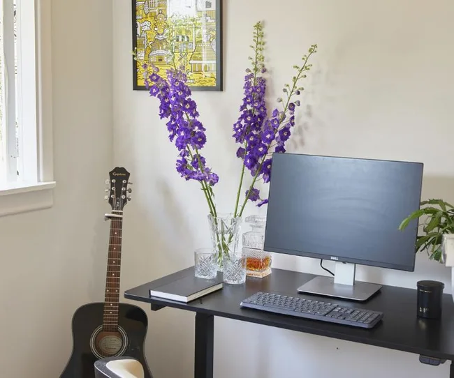 Workspace with a monitor, keyboard, purple flowers in vase, a guitar, and wall art.