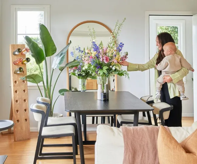 A woman holding a baby arranges flowers on a dining table with modern decor, large mirror, and indoor plants.