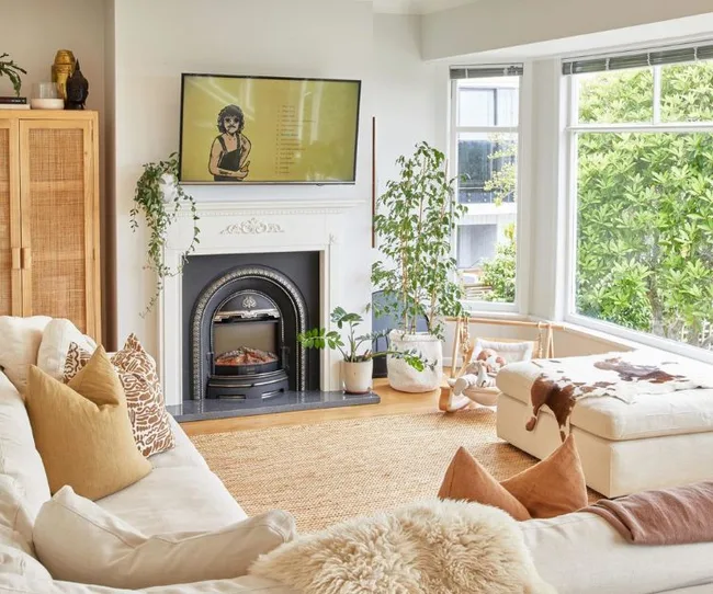 Cozy living room with a TV above a fireplace, beige sofa, and large windows with lush greenery outside.