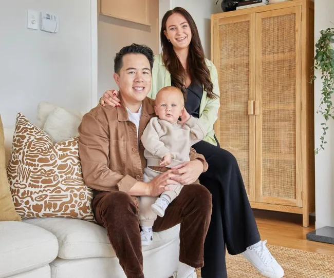A family of three sits on a sofa; a man holds a baby, and a woman stands behind them, smiling.