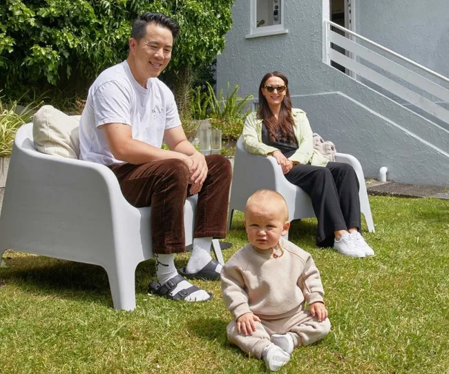 A family sits on lawn chairs, with a baby in the foreground on grass, in a sunny garden setting.