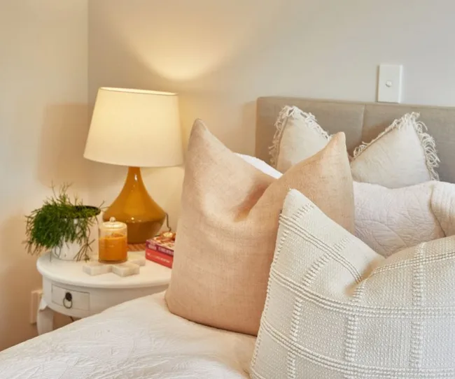 A cozy bedroom with a bed, beige pillows, a lamp, books, a plant, and a candle on a bedside table.