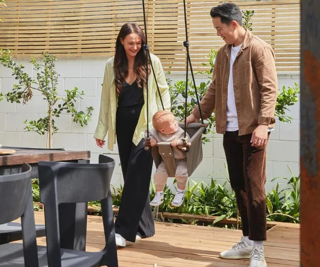 A couple happily pushes a baby in a swing on a sunny deck, surrounded by plants.