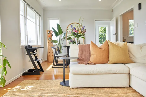 Bright living room with beige sofa, colorful cushions, indoor plants, and a wine rack beside a sunny window.
