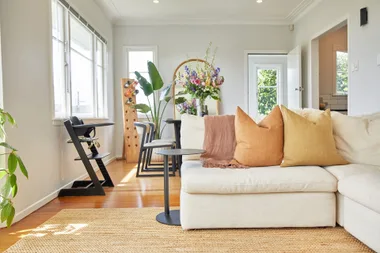 Bright living room with beige sofa, colorful cushions, indoor plants, and a wine rack beside a sunny window.