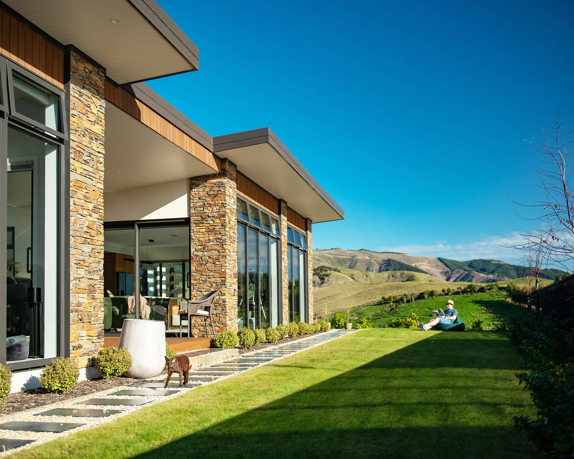 Modern house with stone and glass exterior, dog on patio, person relaxing on lawn, and scenic hills in the background.
