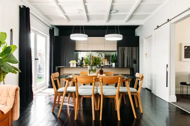 Modern kitchen with a wooden dining table, chairs, pendant lights, and plants, featuring white walls and dark floor.