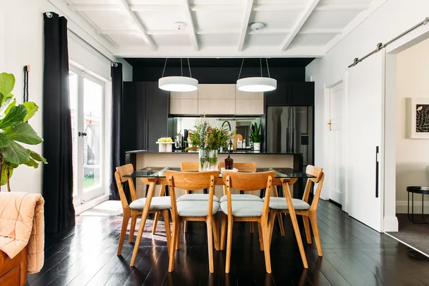 Modern kitchen with a wooden dining table, chairs, pendant lights, and plants, featuring white walls and dark floor.
