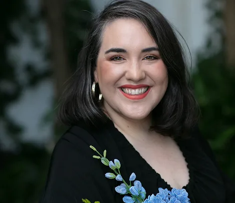 Smiling person with shoulder-length dark hair and hoop earrings, wearing a black top with floral embroidery.