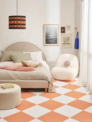 A cozy bedroom with peach checkered floor, bed, armchair, wall art, and a pendant lamp.
