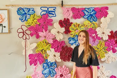 A woman stands in front of a colorful wall of floral yarn art, smiling.