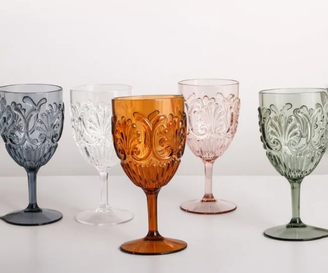 Five ornate colored glass goblets in blue, clear, amber, pink, and green on a white surface.