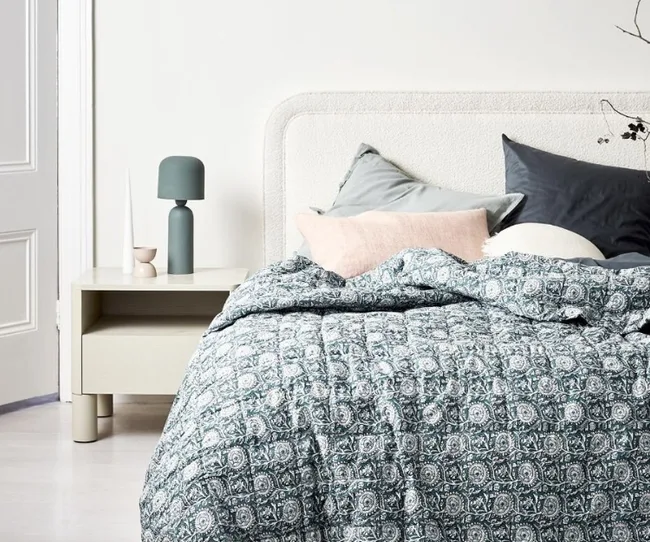 Cozy bedroom with patterned quilt, neutral headboard, decorative pillows, and minimalist side table with lamp.