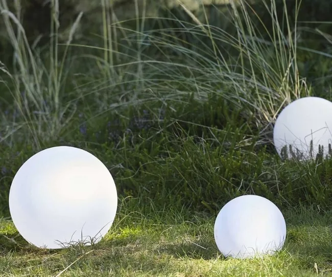 Illuminated round garden lights placed on grass with tall plants in the background.