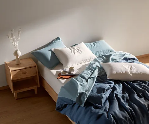 Cozy bedroom with blue and white bedding, a wooden nightstand with a vase, books, and a small cup, in natural light.