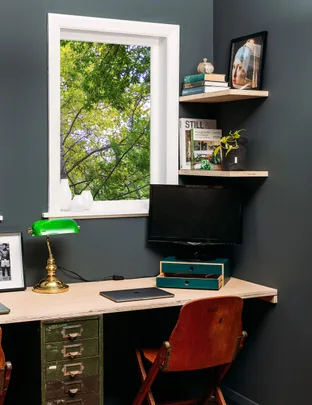 Home office with wooden desk, green lamp, monitor, and shelves. Window view of lush trees.