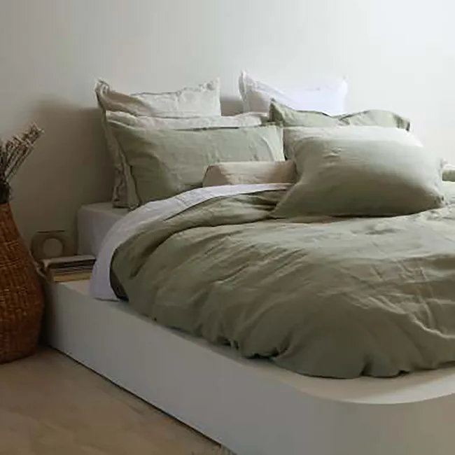 Cozy bed with green linen bedding and pillows, in a minimalist room with a basket and books on the side.