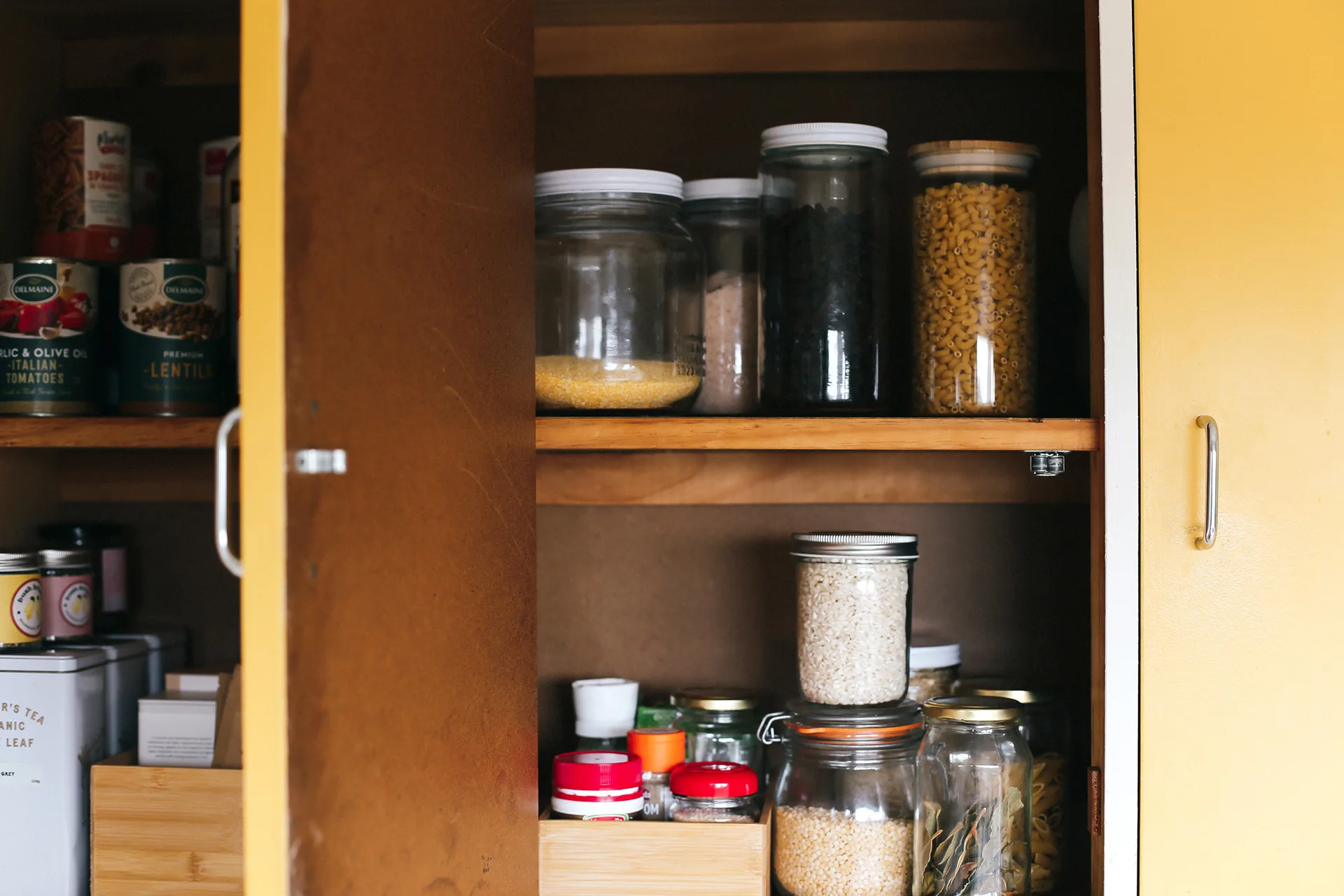 Open kitchen cabinet with jars of grains, beans, pasta, and canned goods neatly arranged on wooden shelves.