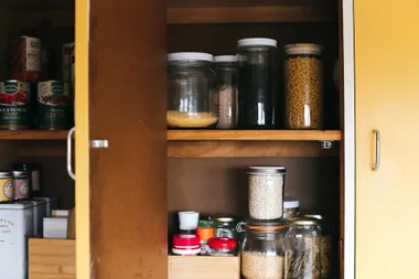 Open kitchen cabinet with jars of grains, beans, pasta, and canned goods neatly arranged on wooden shelves.