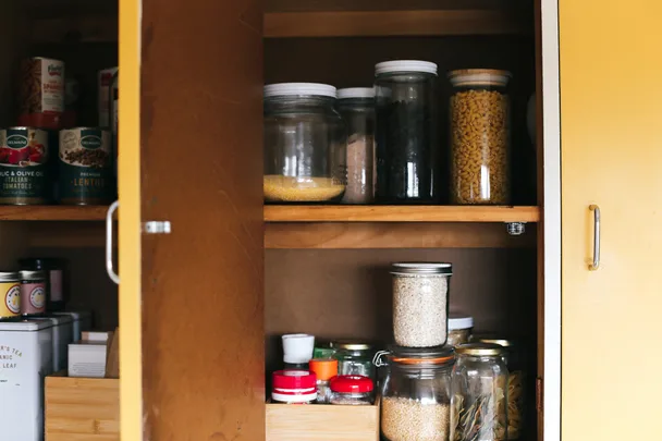 Open kitchen cabinet with jars of grains, beans, pasta, and canned goods neatly arranged on wooden shelves.