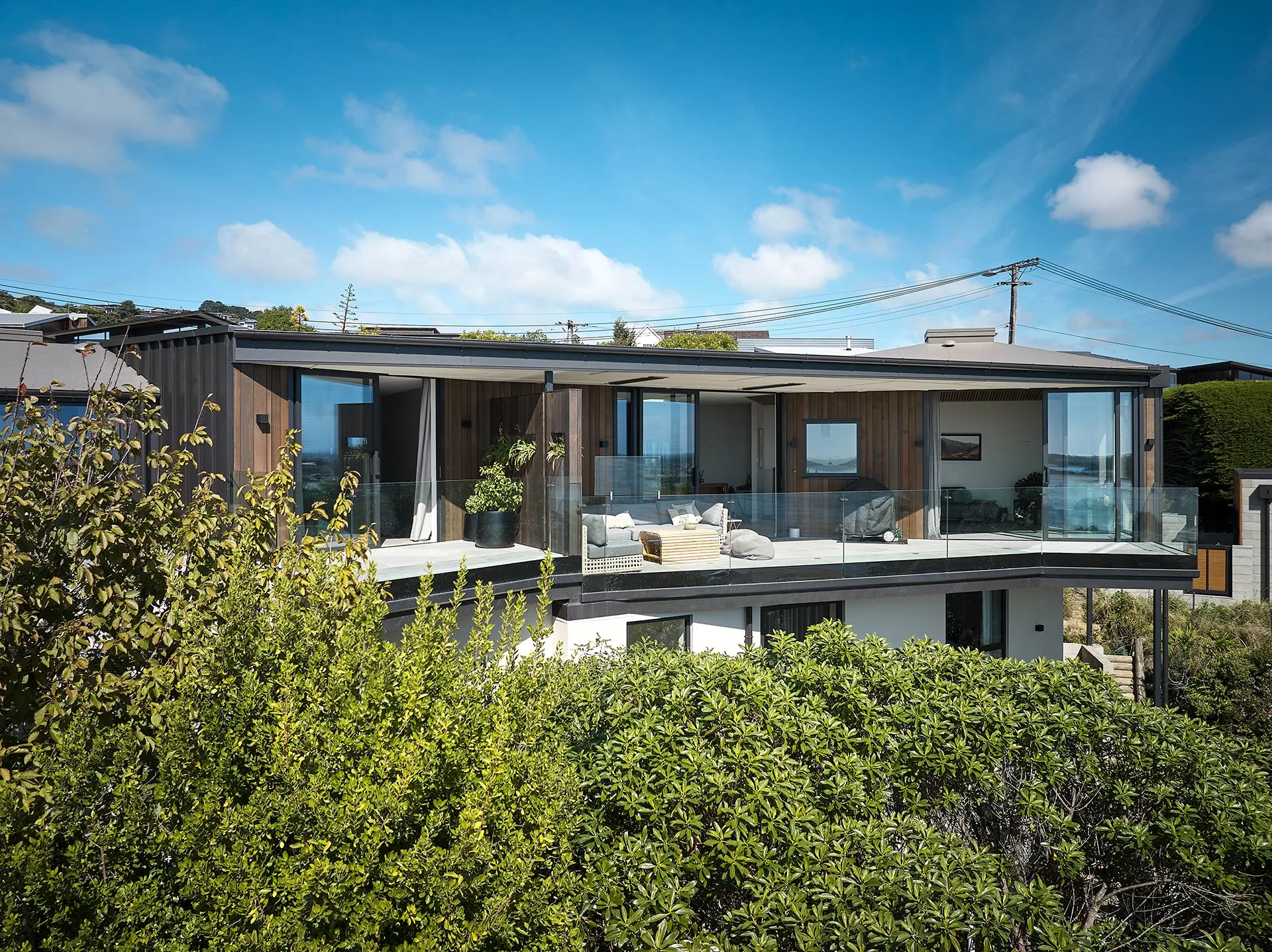 Modern two-story house with large windows and glass balcony, surrounded by lush greenery under a blue sky.