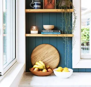 Cozy kitchen nook with blue wall, shelves holding books and decor, and a counter with bowls of fruit and cutting board.