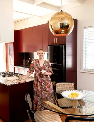 A person in a floral dress stands in a modern kitchen with maroon cabinets, holding a cup next to a round glass table.