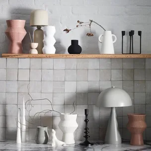 Stylish ceramic vases, candle holders, and lamps on a wooden shelf against a tiled wall.