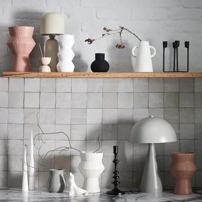 Stylish ceramic vases, candle holders, and lamps on a wooden shelf against a tiled wall.