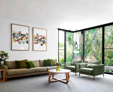 Modern living room with a green sofa, two armchairs, abstract wall art, a round coffee table, and large windows overlooking trees.