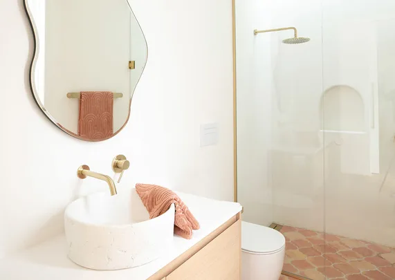 Elegant bathroom with a curved mirror, white basin, gold fixtures, pink towel, and a glass-enclosed shower.