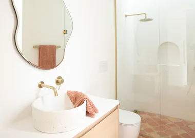 Elegant bathroom with a curved mirror, white basin, gold fixtures, pink towel, and a glass-enclosed shower.
