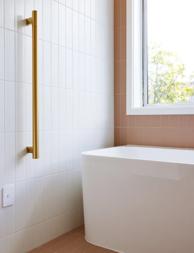 Modern bathroom with white tub, gold towel rail, white and peach tiles, and a window.