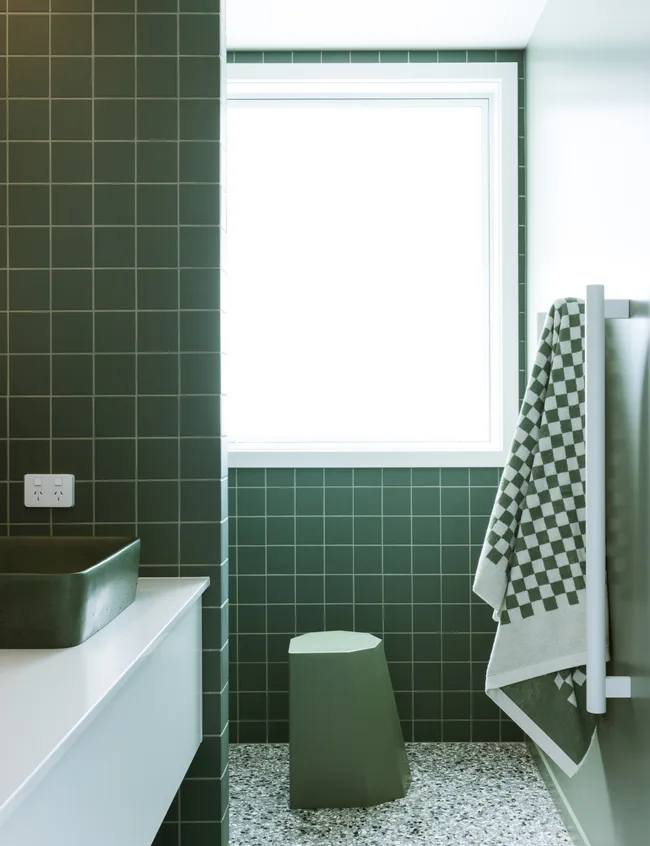 Modern bathroom with green tiles, a white sink, geometric stool, and a checkered towel on a rack near the window.