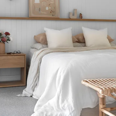 Cozy bedroom with a white bedspread, wooden nightstand, flowers, glasses, and minimal decor.