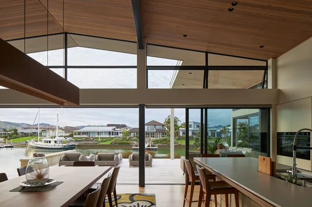 Modern living room with large windows overlooking a canal and houses, featuring wooden ceiling and dining area.