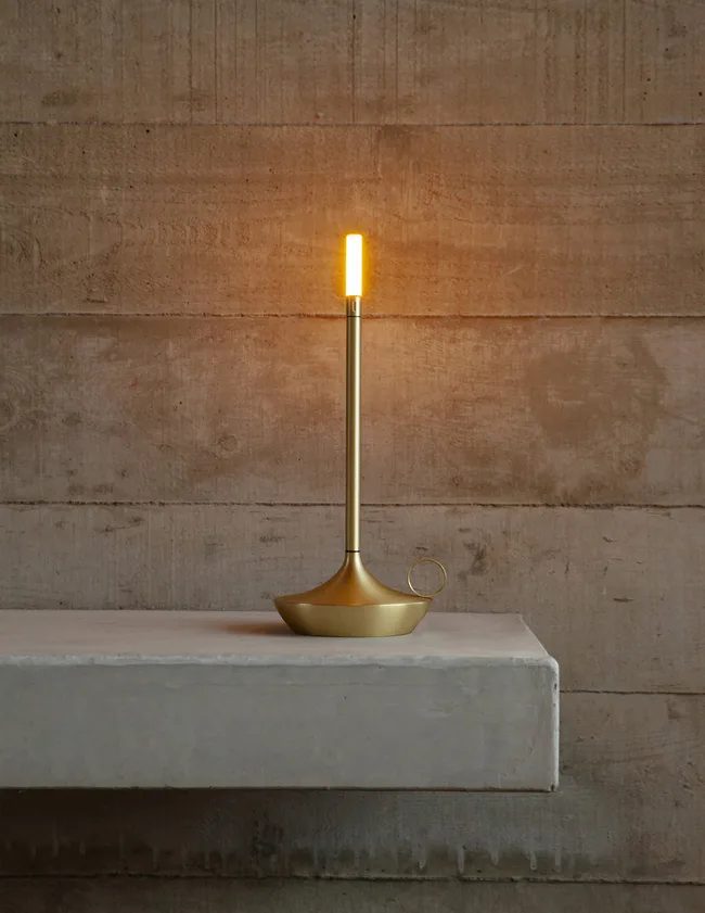 Golden modern candle-shaped lamp on concrete shelf against textured wall.
