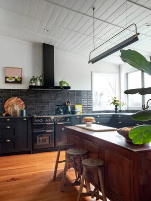 A modern kitchen with black cabinets, wooden island, stools, and plants, featuring a window and hanging light fixture.