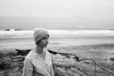 A person wearing a knit hat and sweater stands on a beach, gazing out at the ocean in a black and white photo.