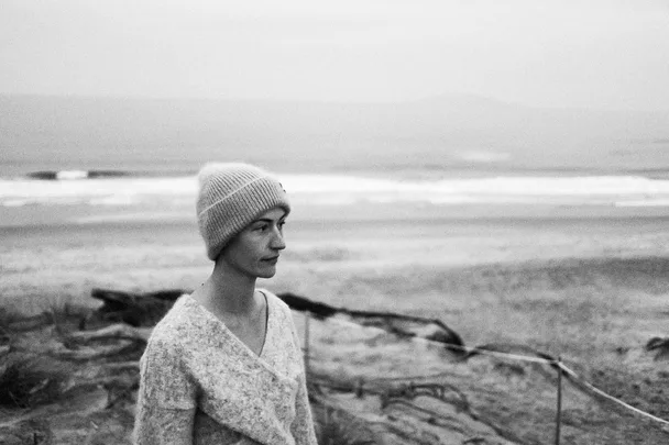 A person wearing a knit hat and sweater stands on a beach, gazing out at the ocean in a black and white photo.