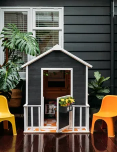 A small black and white playhouse with a plant on its door step, flanked by two yellow chairs and large leaves.