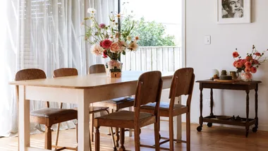 Dining room with wooden table, chairs, flowers in vases, two green pendant lights, and a monochrome portrait on the wall.