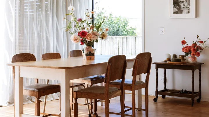 Dining room with wooden table, chairs, flowers in vases, two green pendant lights, and a monochrome portrait on the wall.