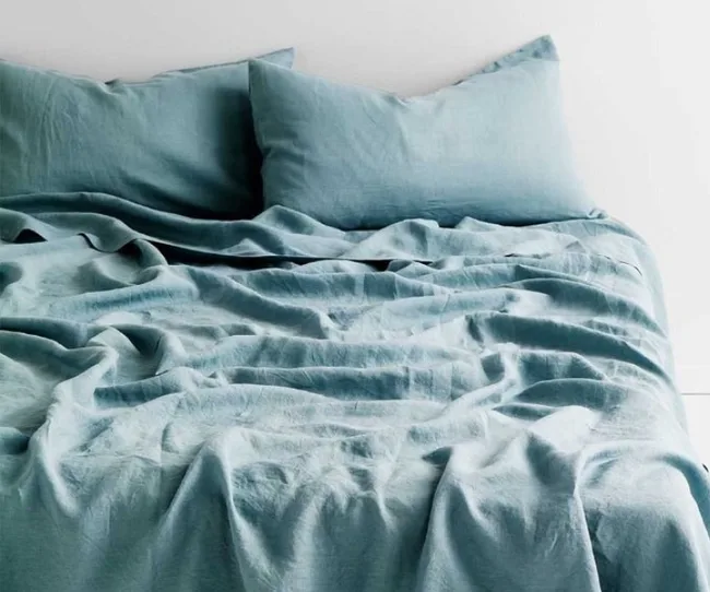 Unmade bed with crumpled teal sheets and two matching pillows against a white wall.