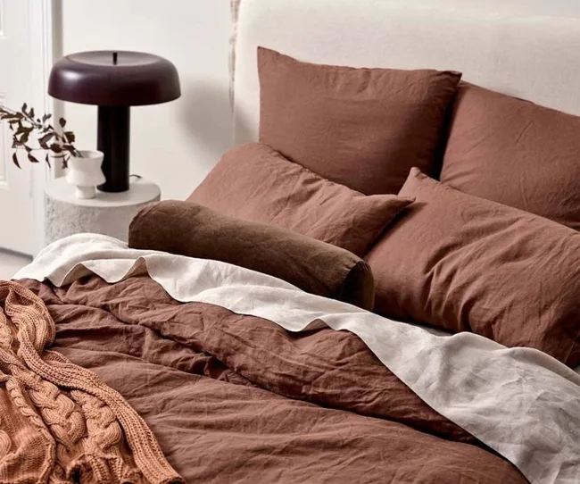 A neatly made bed with brown bedding, a textured blanket, and a round pillow; dark lamp on a side table with decor.
