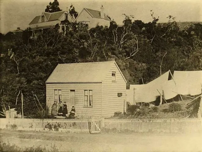 Historic sepia photo of a small wooden house with a picket fence, people at the door, and tents in the background.